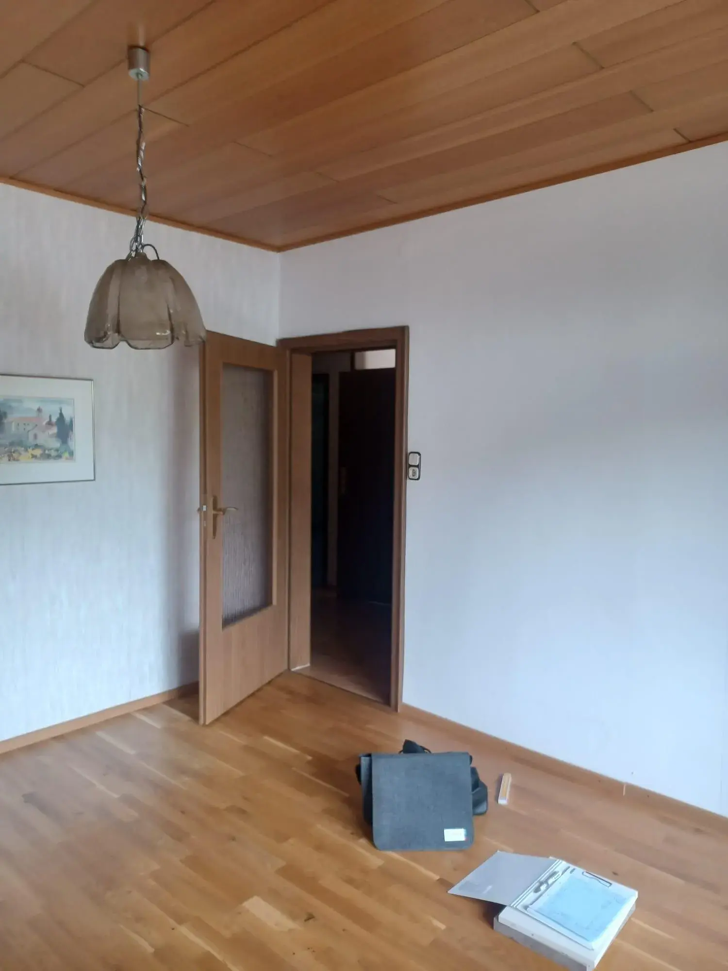 Leeres Zimmer mit Holzboden, offener Tür, Lampe und Ordner auf dem Boden.